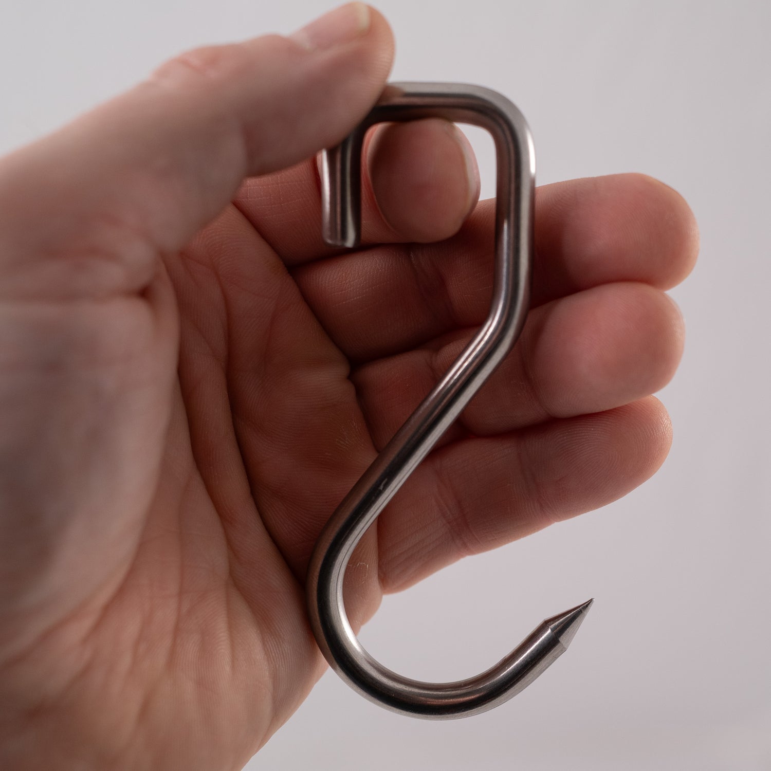Hand holding a metallic s-hook, a food safe sharp stainless steel meat hook on a white background
