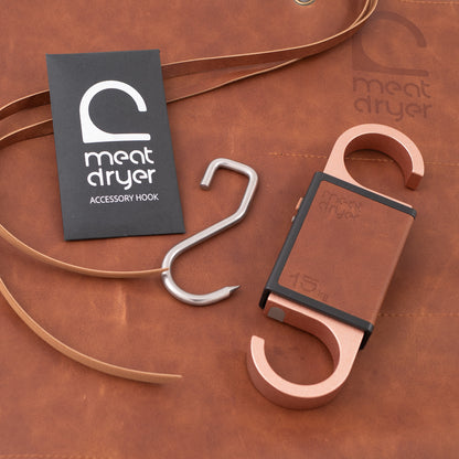 Two hooks, one sharp steel metal accessory hook and one Meatdryer Artisan smart meat hook, on a brown leather surface with &